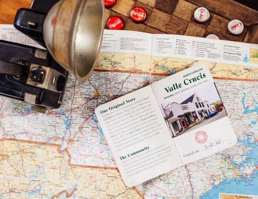 A vintage-style open passport showing the Original Mast Store Valle Crucis location laid on top of a detailed map, surrounded by a black camera with old-fashioned flash reflector, and Coca-Cola bottle caps on a wooden checkerboard.