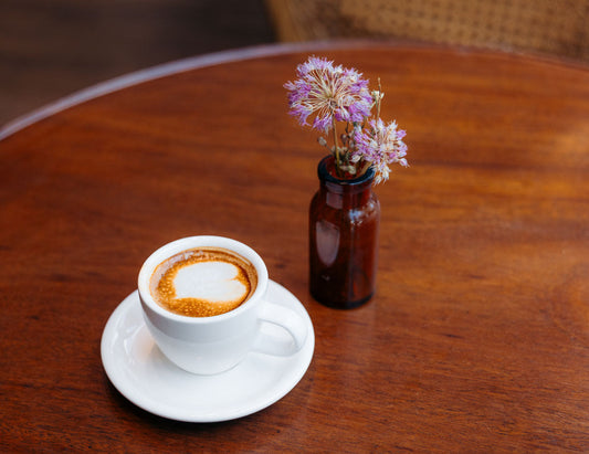 A latte in a white cup has a heart shape poured on top sitting on a medium brown colored round wooden table sitting next to a small brown vase with purple flowers.