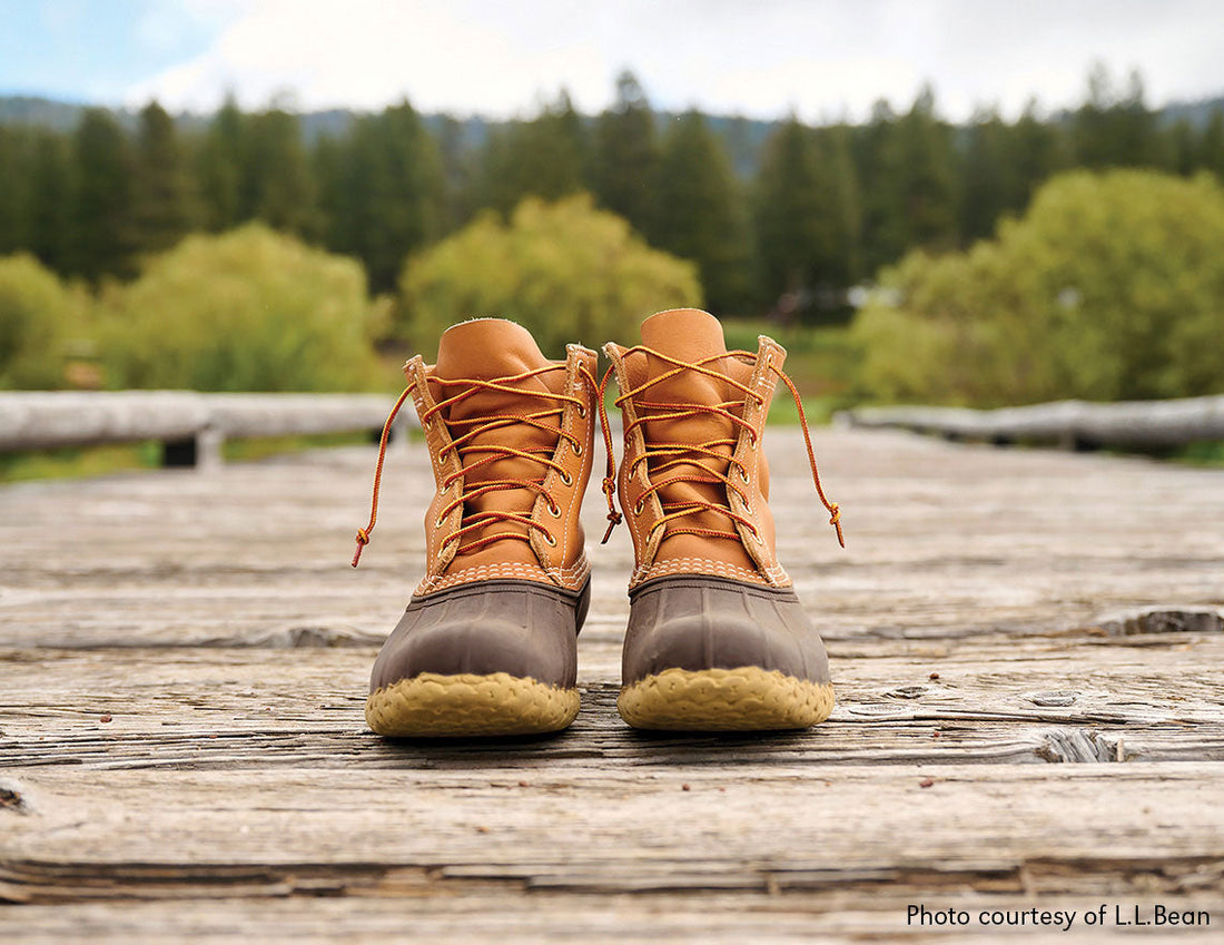 A pair of boots with leather uppers and rubberized toe box area sitting on an old wooden dock with a blurred forested background. These boots are Made in USA by L.L.Bean.
