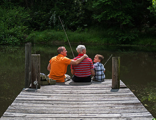 Three generations of men fishing on a dock at a pond surrounded by green foliage.