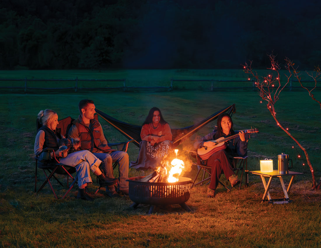 Four people sitting around a campfire at night in an outdoor grassy field, with two more people lounging in a hammock behind them. One person holds a guitar, and a small side table with a lit candle stands to the right.