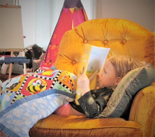 Small child in a chair reading while covered in a childrens' blanket.