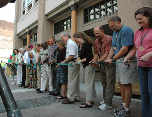 A group of people standing in a line outdoors, cutting a green ribbon at what appears to be a ceremonial opening in front of a brick building.
