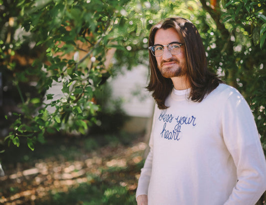 Man with long brown hair and glasses wearing a long-sleeved white shirt with bless your heart on the front stands in a forested area.