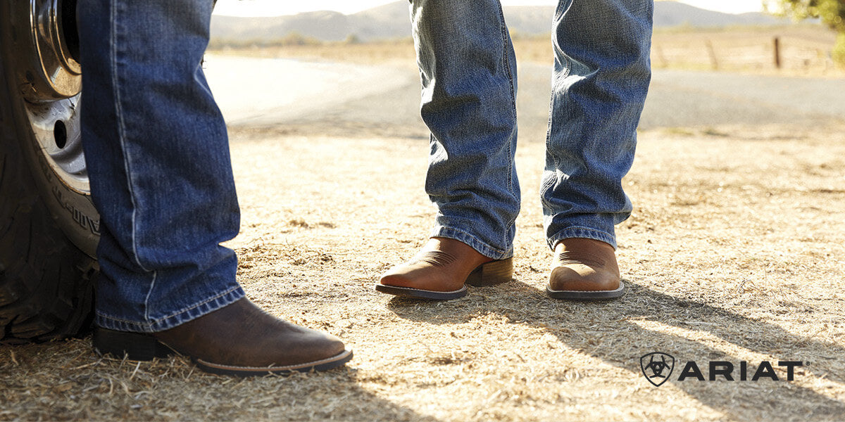 Close-up of two people wearing blue jeans and brown Ariat western-style boots standing on a dirt road