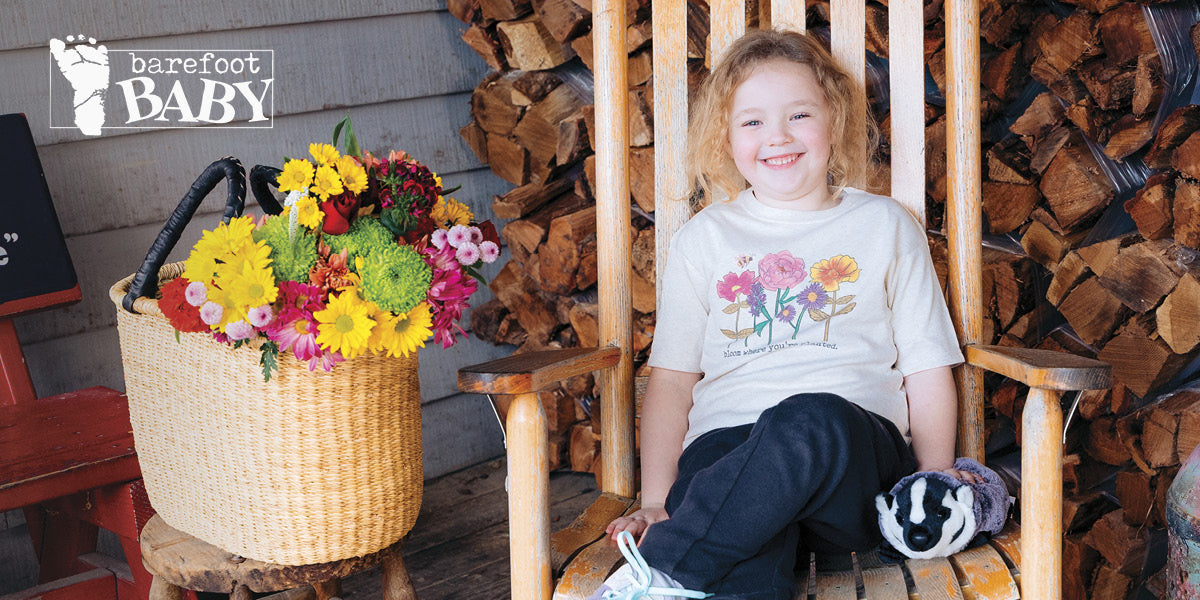 Girl in a Barefoot Baby tee sitting on a rocking chair next to a basket of flowers.