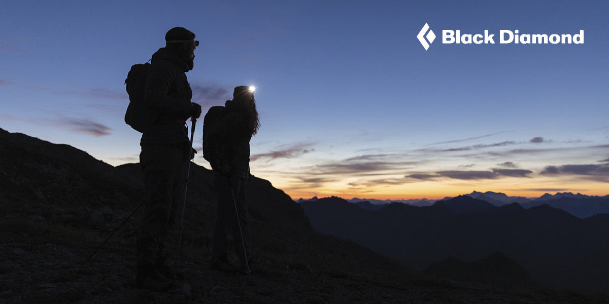 Two hikers silhouetted wearing Black Diamond headlamps against a mountainous landscape with a twilight sky.