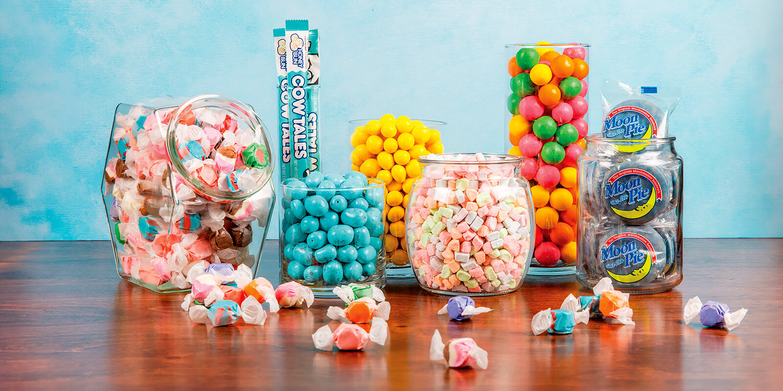 Assorted colorful candies in jars and bags on a wooden surface with a blue gradient background