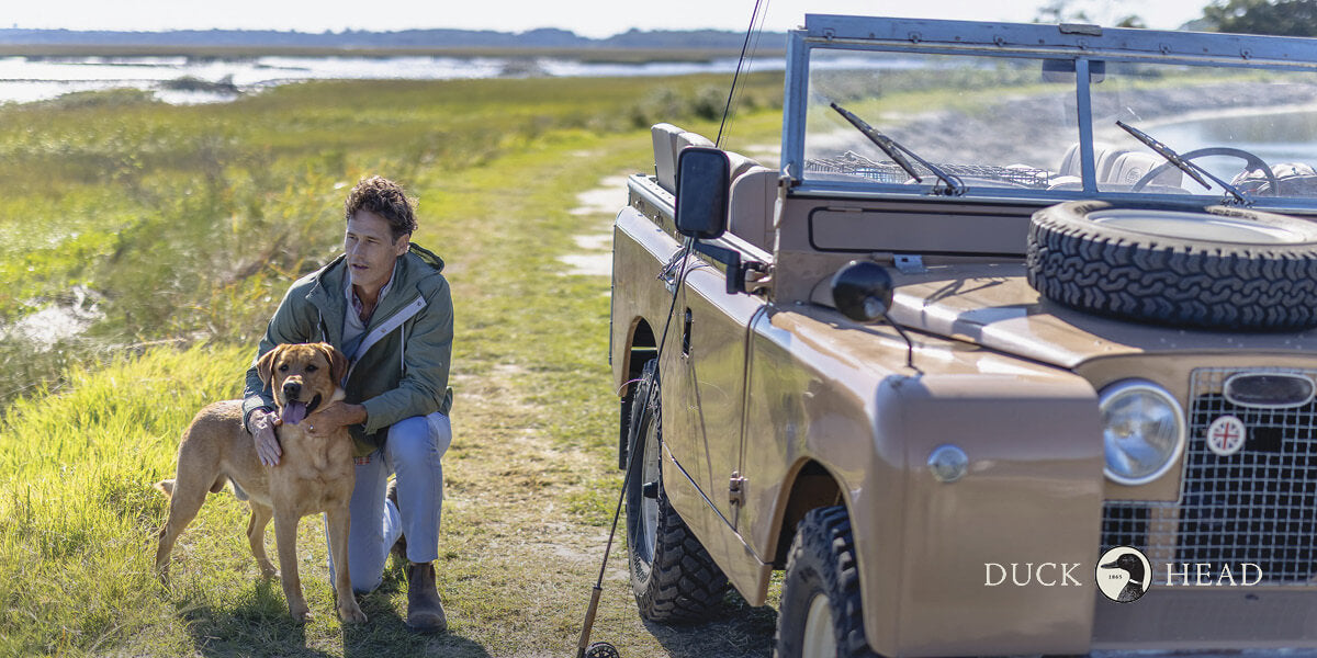 A man in a Duck Head top patting a dog next to a vintage Land Rover.