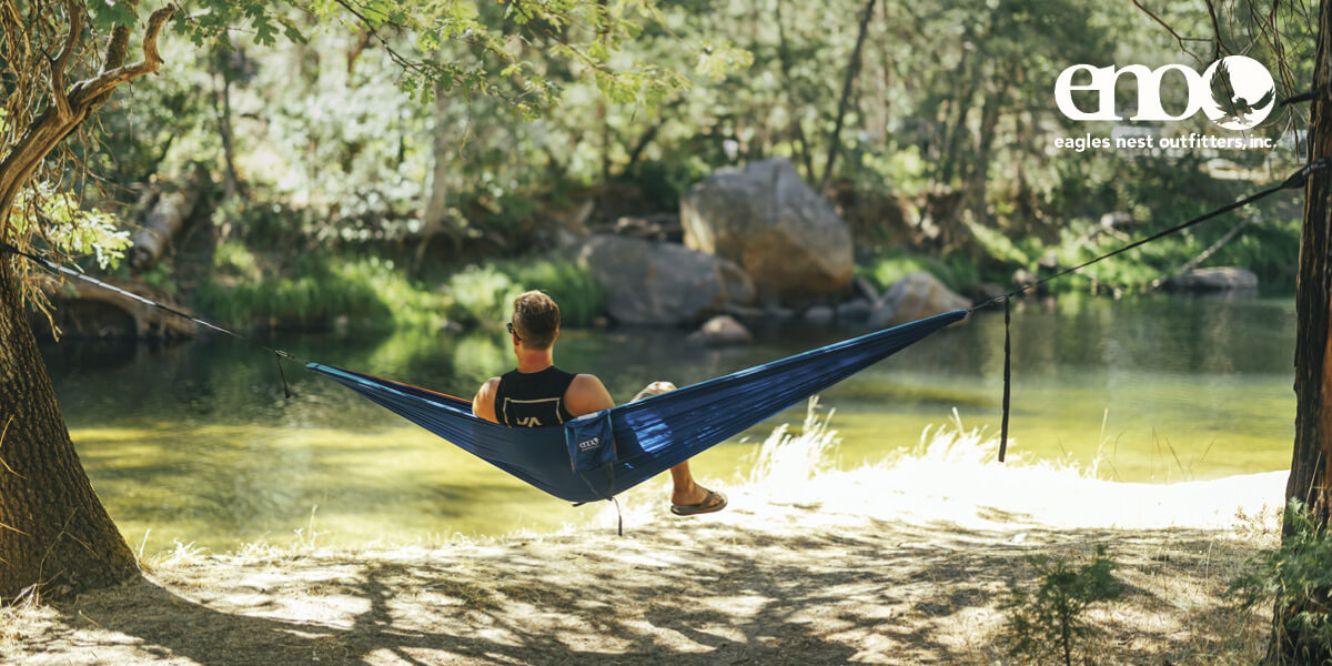 Person relaxing in a ENO hammock by a river surrounded by trees.