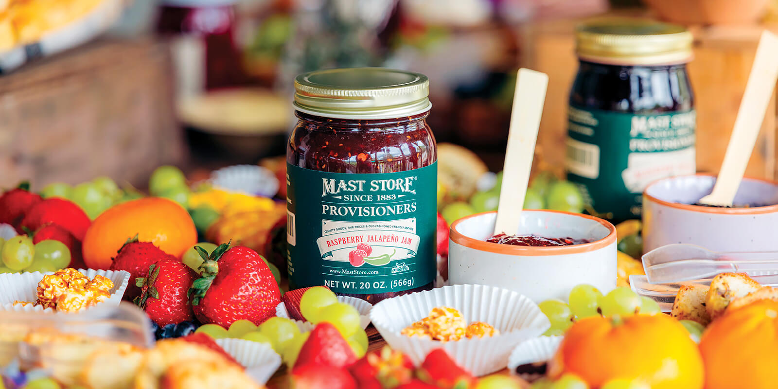 A Mast Store Provisioners Raspberry Jalapeño Jam jar displayed on an event food table with colorful fresh fruits and other food items softly out of focus