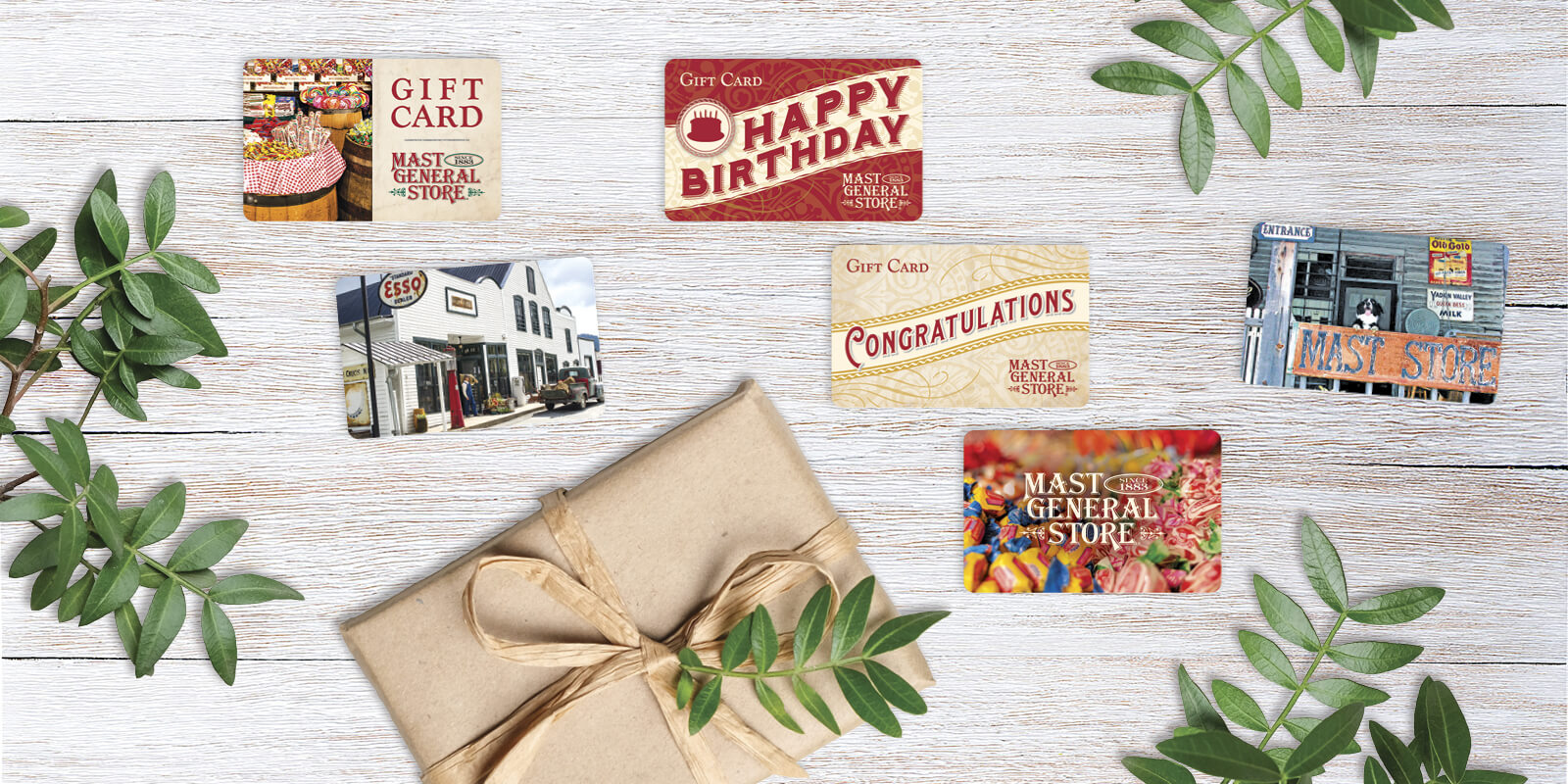 A collection of six gift cards, including birthday, congratulations, and Candy Barrel designs, displayed beside a kraft paper–wrapped gift on an off‑white wooden surface with greenery