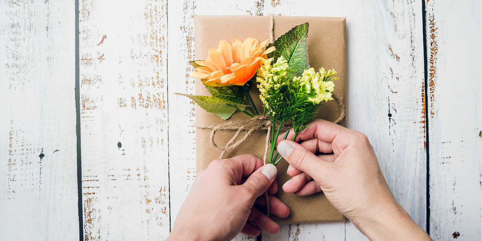 A person tying a bouquet of flowers onto a wrapped gift on an off‑white wooden surface
