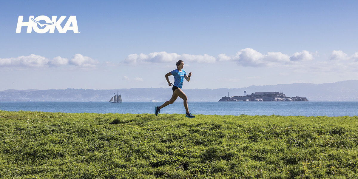 Person running on grass with a scenic view of water and an island in the background, featuring Hoka shoes.