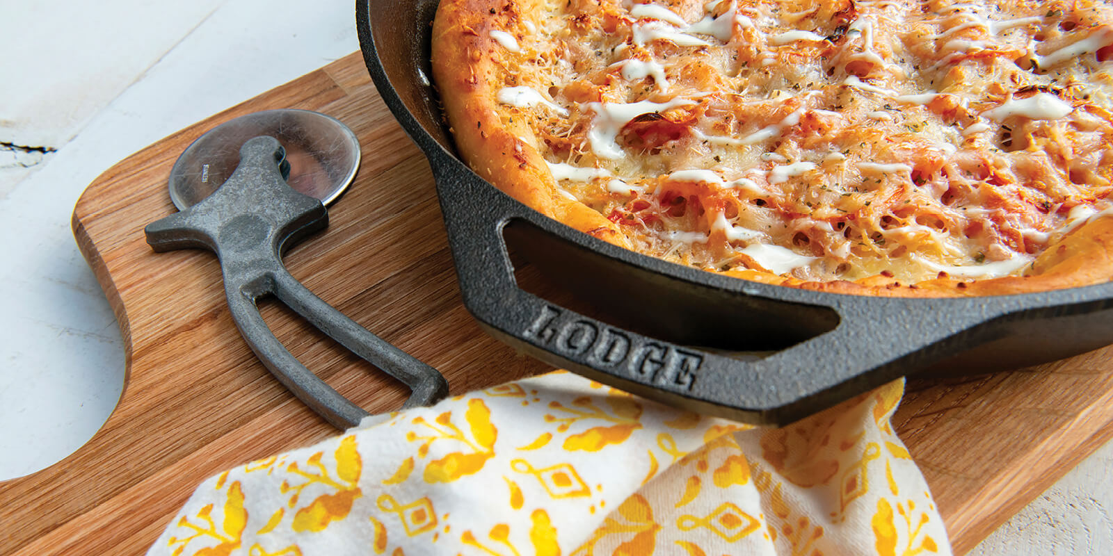 A freshly baked homemade pizza in a Lodge cast iron Baker’s Skillet placed on a wooden board with a pizza cutter and a kitchen towel