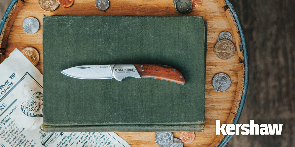 Folding Kershaw knife resting on a green book with coins on a wooden barrel.