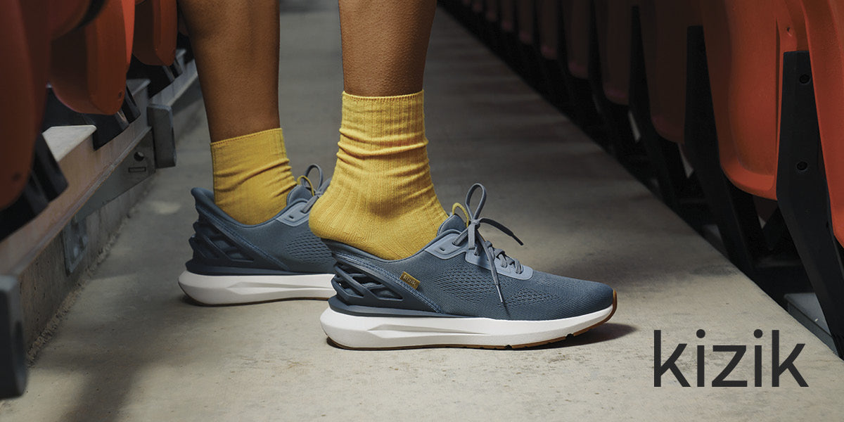 Person putting on blue Kizik sneakers with yellow socks on a concrete floor.
