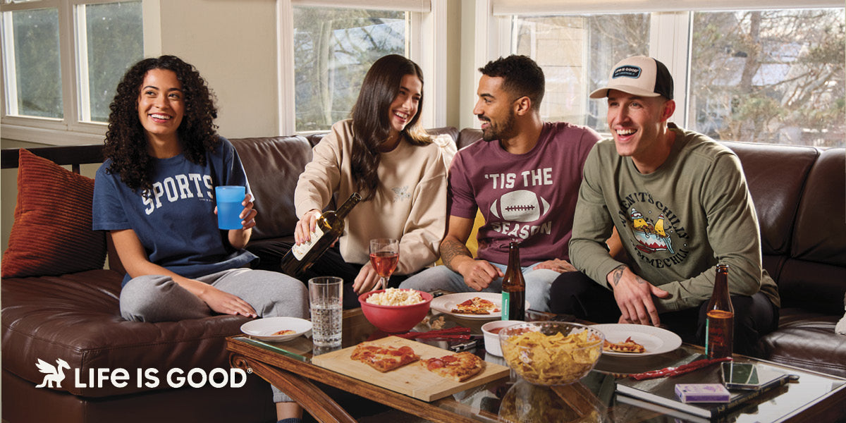 Four friends sitting on a couch, enjoying pizza and drinks together wearing Life is Good tees.