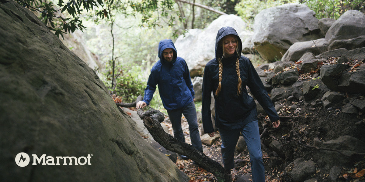 Two hikers wearing Marmot rain jackets walking through a rocky forest trail.