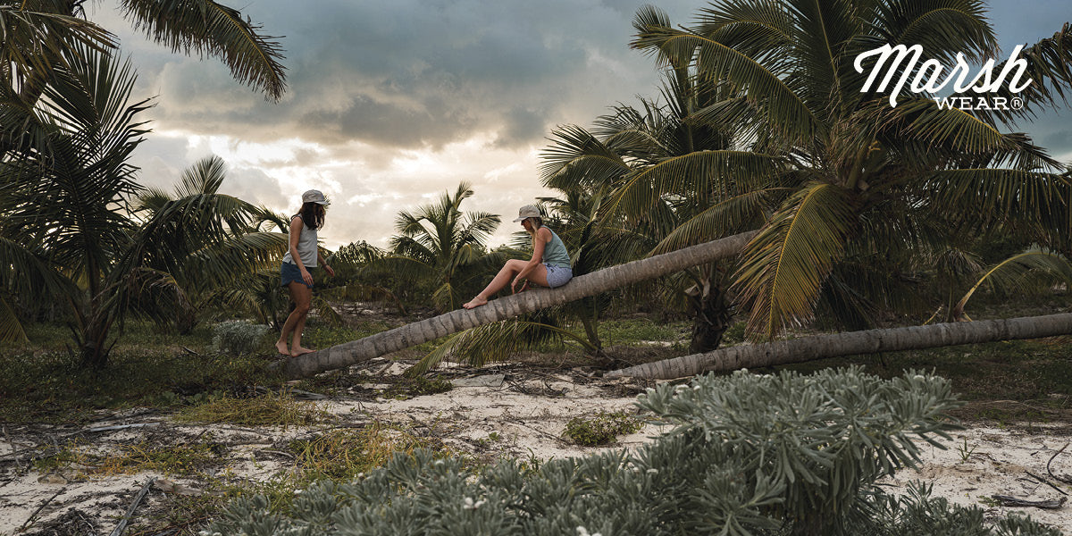 Two people sitting on a fallen palm tree in a tropical setting, wearing Marsh Wear apparel.