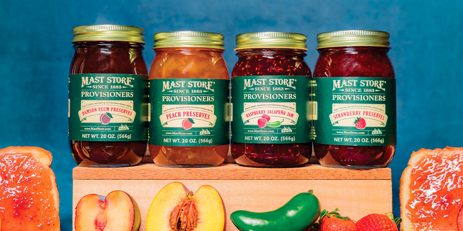 Four jars of Mast Store Provisioners fruit preserves displayed on a wooden stand, with fresh‑cut fruit in the foreground and two slices of bread spread with preserves against a blue background