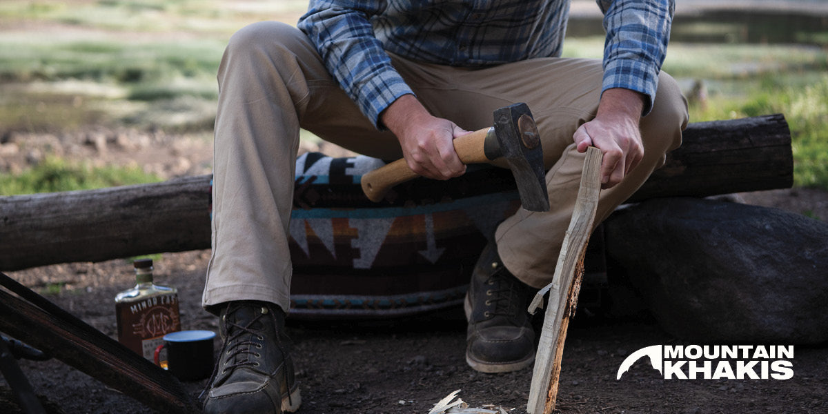 Person sitting on a log with an axe, wearing Mountain Khakis clothing, in an outdoor setting.