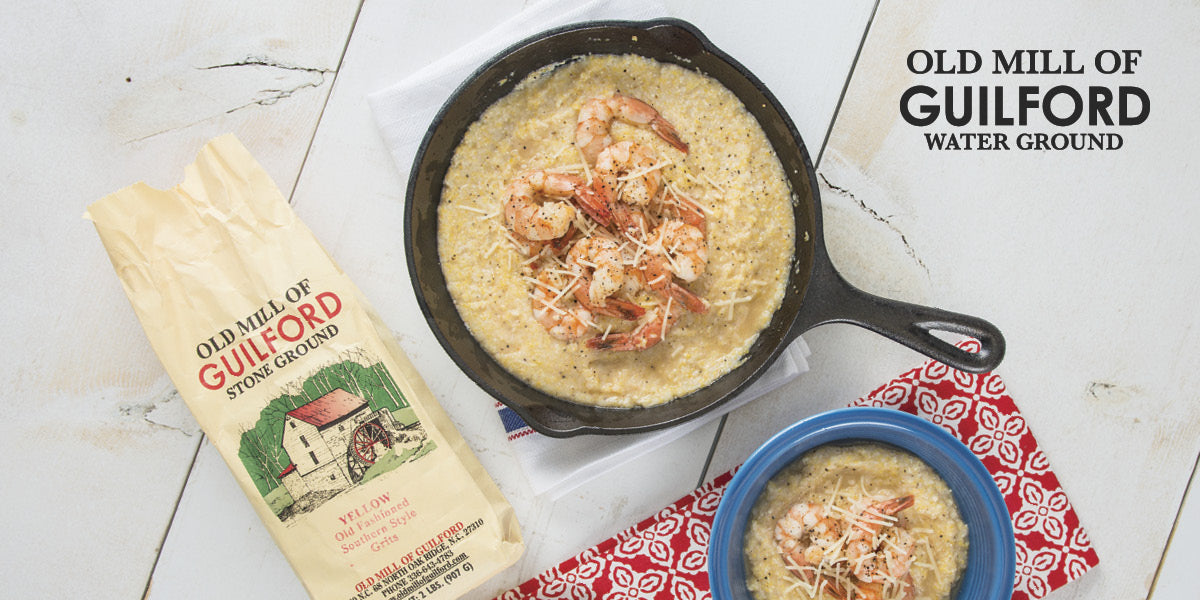 Shrimp and grits in a skillet, next to a pack of Old Mill of Guilford grits on a white wooden surface.