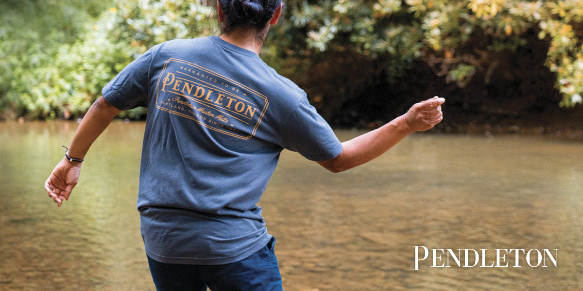 Person wearing a Pendleton shirt tossing a rock into a river.