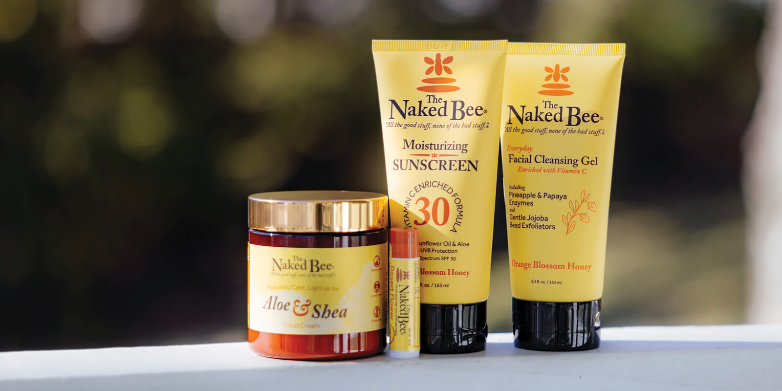 The Naked Bee skincare products, including Aloe & Shea Cloud Cream, Moisturizing Sunscreen, and Facial Cleansing Gel, displayed against a blurred sunny outdoor background