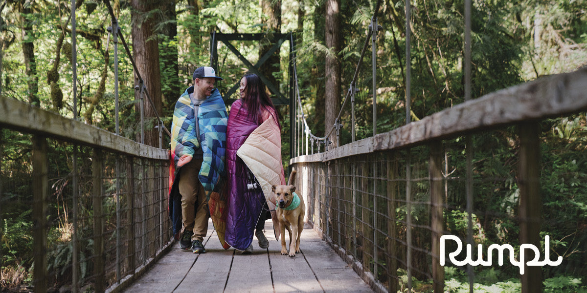 Two people walking a dog on a wooden bridge in a forest, wrapped in colorful Rumpl blankets.