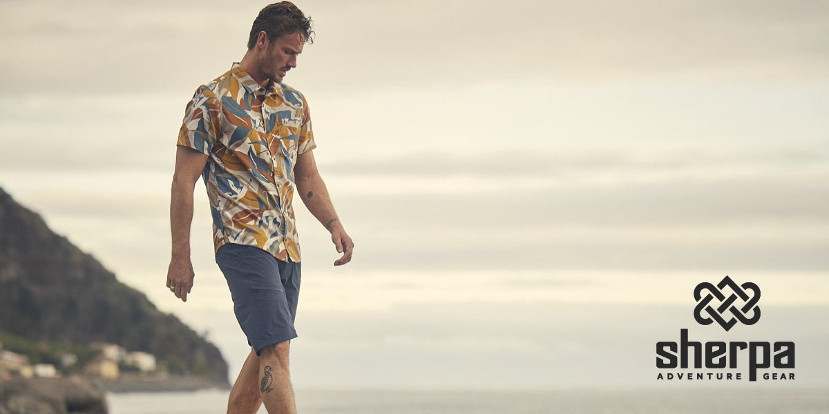 Man walking along a beach with mountains in the background, wearing a Sherpa Adventure Gear patterned shirt and shorts.