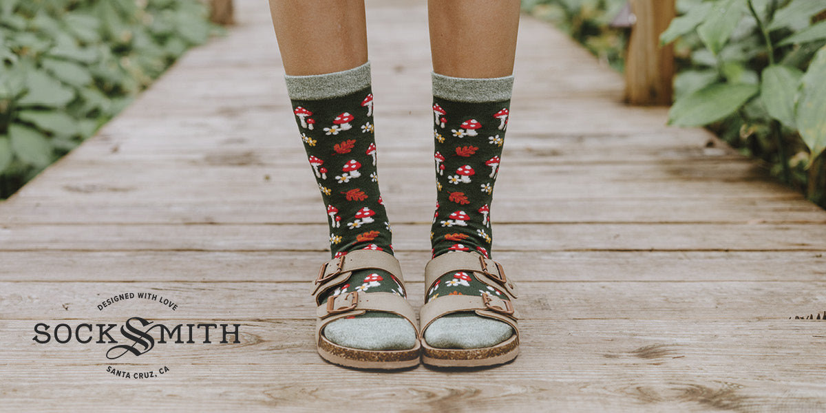 Person wearing Socksmith mushroom‑patterned socks with sandals on a wooden path.