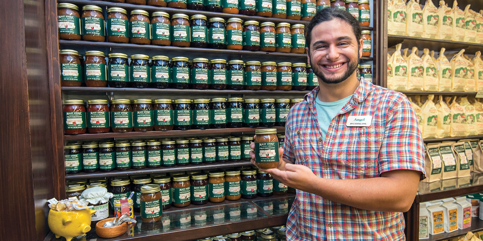 A Mast Store employee holding a Mast Store Provisioners jar of jam in front of shelves stocked with Mast Store Provisioners jams, jellies, and preserves inside the store