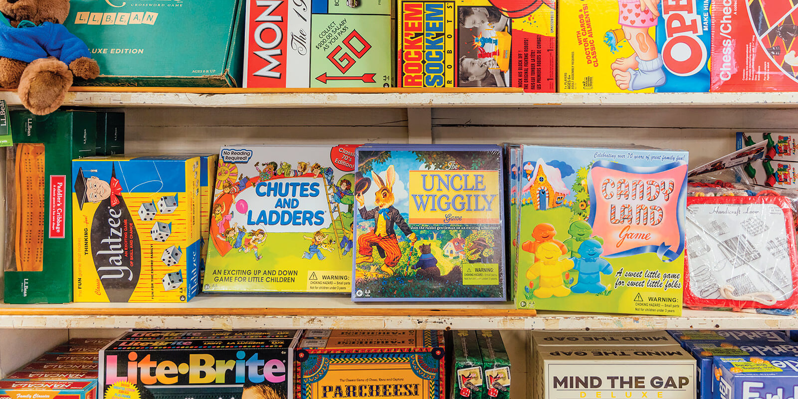 A shelf in the Mast General Store toy department displaying board games and craft toys, including L.L.Bean Paddler’s Cribbage, Yahtzee, Candy Land, and Weaving Loops and Loom Craft