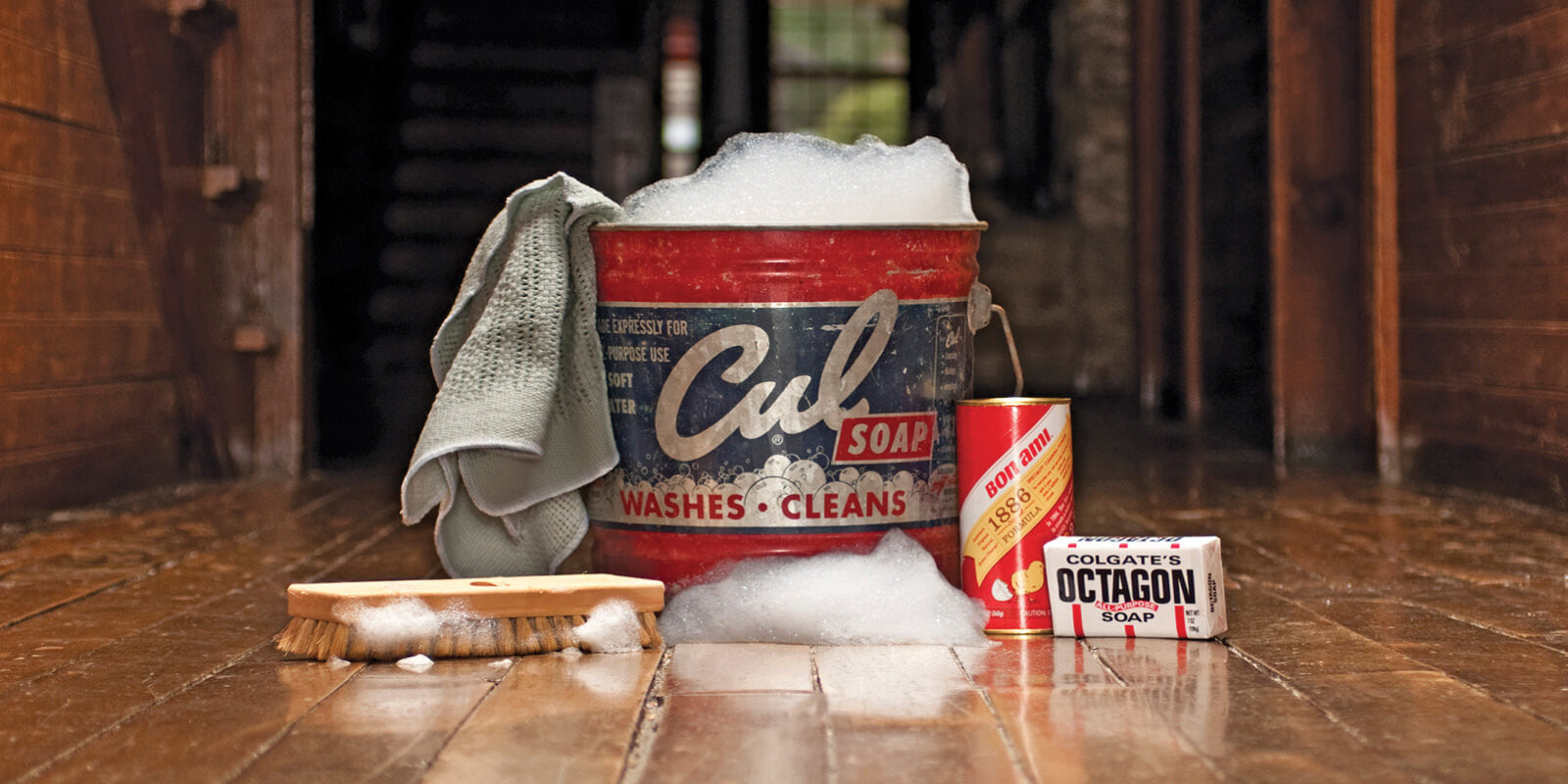 Vintage‑style cleaning supplies, including a bucket labeled “Cul Soap,” a cleaning rag, a brush, Bon Ami Powder Cleanser, and a bar of Colgate’s Octagon All‑Purpose Soap, arranged on a wooden floor