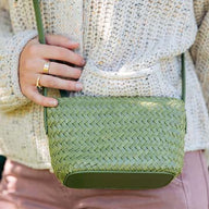 A person wearing a cream knit sweater and pink pants holds a green woven crossbody bag with a matching strap.