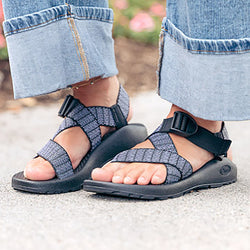Close-up of a person wearing blue and white patterned Chaco sandals with rolled-up blue jeans, standing on a paved outdoor surface