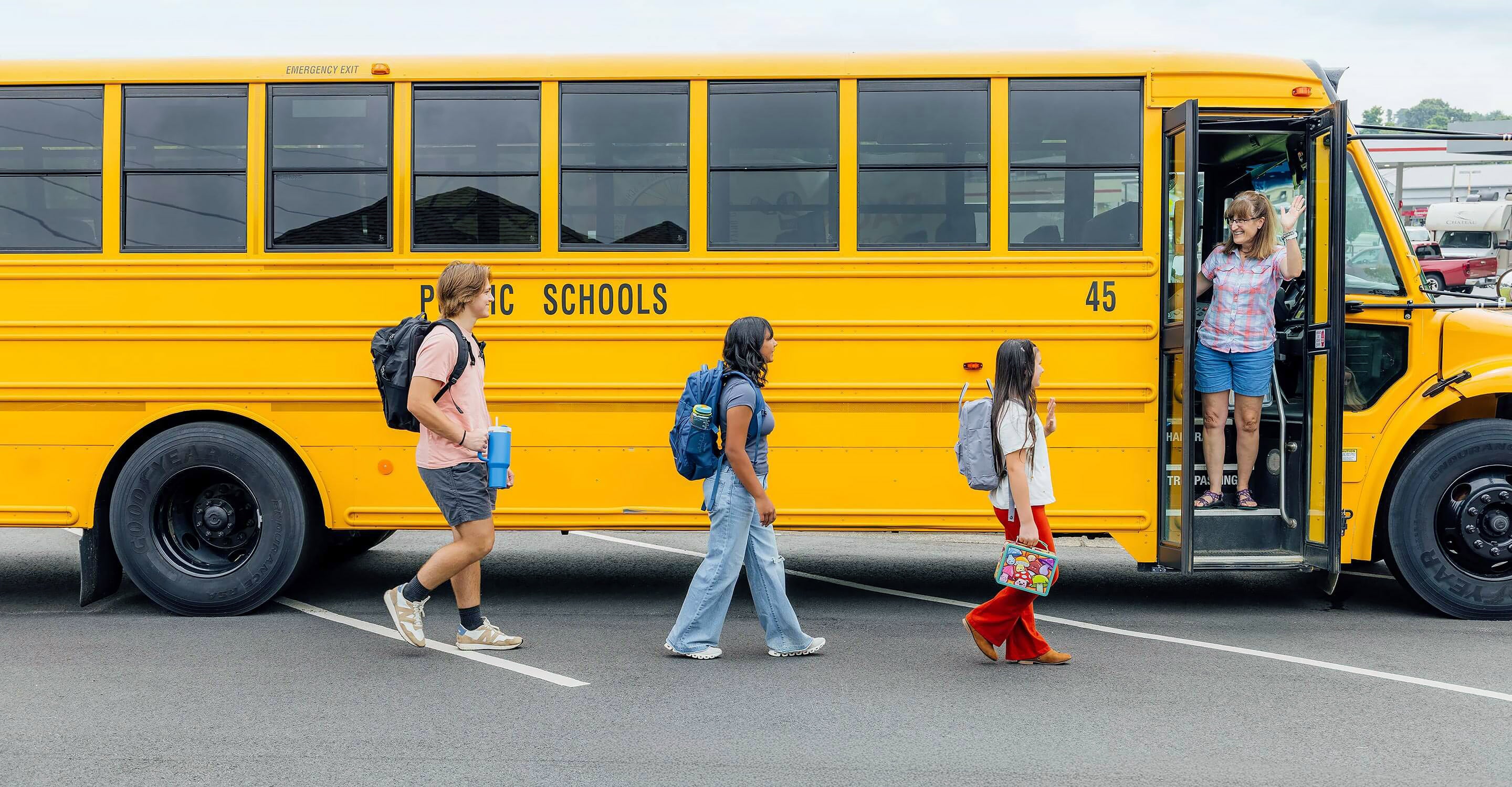 A school bus driver greets three children as they approach the school bus with its door open in a paved parking area. Vehicles and buildings are visible in the background