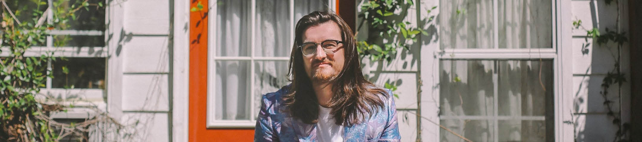 Man, Landon Bryant, with long hair and glasses standing in front of a white house and a door painted orange.