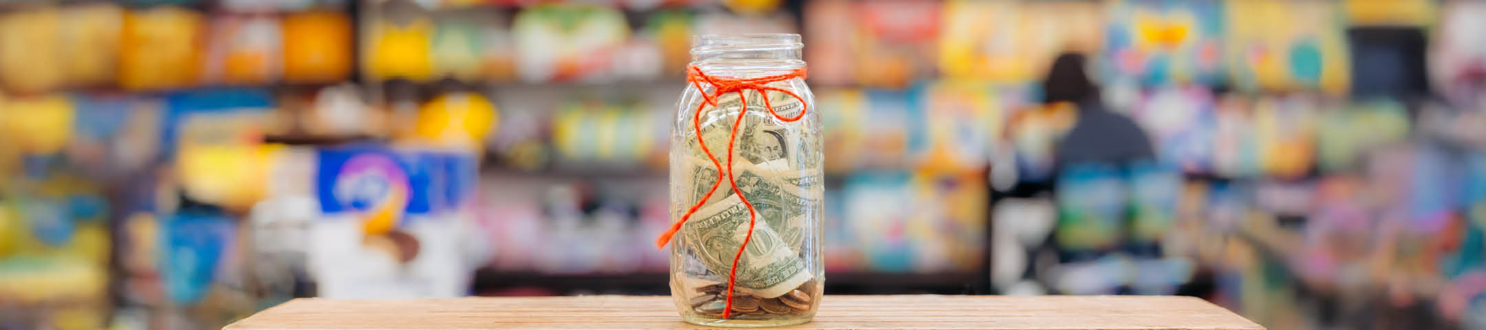 Jar of money with a red string tied around it on a counter with a blurred store background