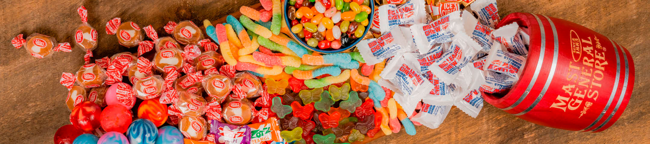 Assorted colorful candies including gummy bears and wrapped Bullseyes on a wooden surface with a mini red barrel.