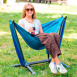 Woman sitting in a blue hammock chair in a park, holding a drink.